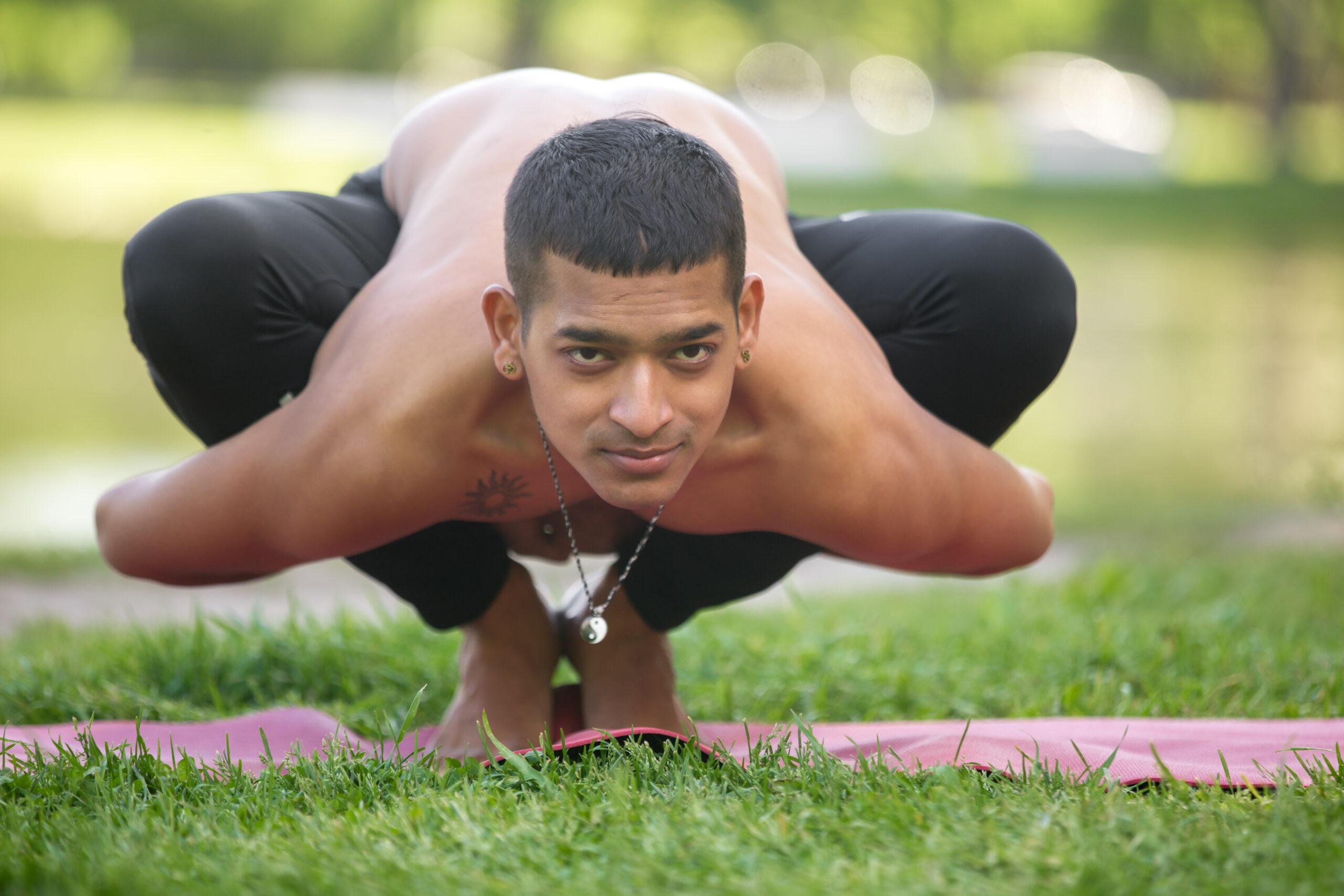 Attractive sporty Indian young yogi man working out on lakeside in park, doing yoga, fitness or pilates training, sitting in squat, Garland Pose, Malasana